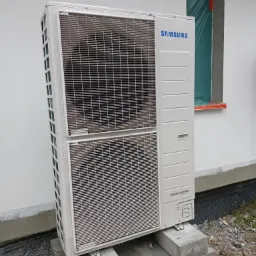 Biała, zewnętrzna jednostka klimatyzacji Samsung Digital Inverter, osadzona na betonowych blokach przy ścianie budynku z oknem zabezpieczonym folią budowlaną, widoczne ślady wilgoci na obudowie.