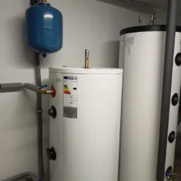 Biały, cylindryczny zbiornik na wodę Soltank 200 z etykietą energetyczną C, obok drugi zbiornik z izolacją, niebieskie naczynie wzbiorcze z manometrem na ścianie, szare rury z zaworami.