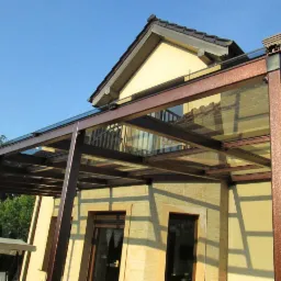 Zadaszenie tarasu z grafitowym szkłem, wsparte na metalowej konstrukcji w kolorze miedzianym, przylegające do jasnej ściany budynku z widocznymi oknami i fragmentem dachu.