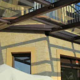 Zadaszenie ze szkłem grafitowym nad oknami w elewacji budynku z widocznym fragmentem balkonu na wyższej kondygnacji.