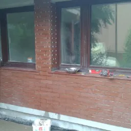 Montaż klinkieru na elewacji zewnętrznej domu, widoczne okna z brązowymi ramami, przymocowane płytki z krzyżykami dystansowymi, narzędzia pracy na parapecie i wiadro z zaprawą na dole.