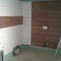 Wnętrze w trakcie prac wykończeniowych: częściowo ułożona pozioma okładzina imitująca drewno na płycie kartonowo-gipsowej, widoczne otwory na instalacje, zielona lamperia, skrzynka z narzędziami...