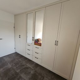 Artex Kuchnie i Wnętrza Natalia Kubiak - Biała szafa wnękowa z lustrami i czarnymi uchwytami, wkomponowana w jasne wnętrze z ciemną podłogą. Widoczny fragment pokoju z drzwiami.