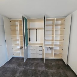 Artex Kuchnie i Wnętrza Natalia Kubiak - Biała, zabudowana szafa na wymiar z otwartymi drzwiami, widoczne półki i szuflady, metalowe wieszaki na spodnie, ciemna podłoga, minimalistyczny design.