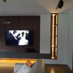 Nowoczesna szafa wnękowa z białymi frontami i podświetlanymi półkami, wkomponowana w aranżację salonu z telewizorem i drewnianą ścianą.