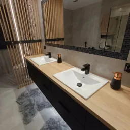 Nowoczesna łazienka z drewnianym blatem, dwiema białymi umywalkami z czarnymi bateriami, podświetlaną szafką pod umywalką i dekoracyjną ścianką z drewnianych listew.