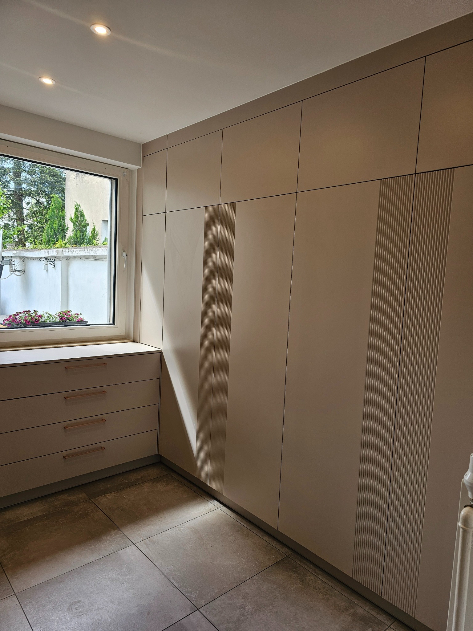 Beżowa szafa wnękowa z ryflowanymi panelami, dopasowana do okna z widokiem na zielony ogród, na tle płytek podłogowych w odcieniach szarości.