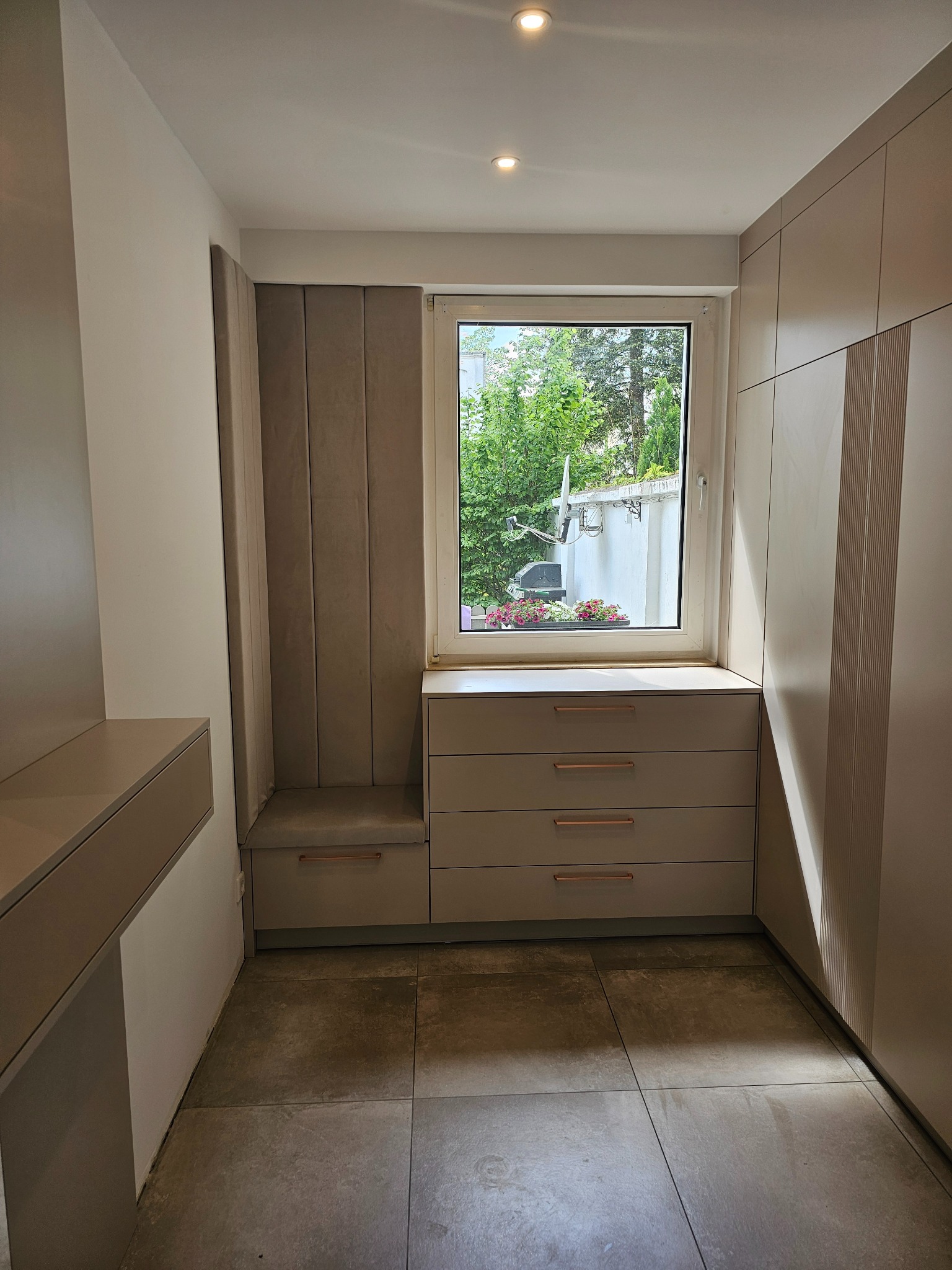 Wnętrze garderoby z beżowymi szafami na wymiar, siedziskiem obok okna i szufladami z miedzianymi uchwytami.