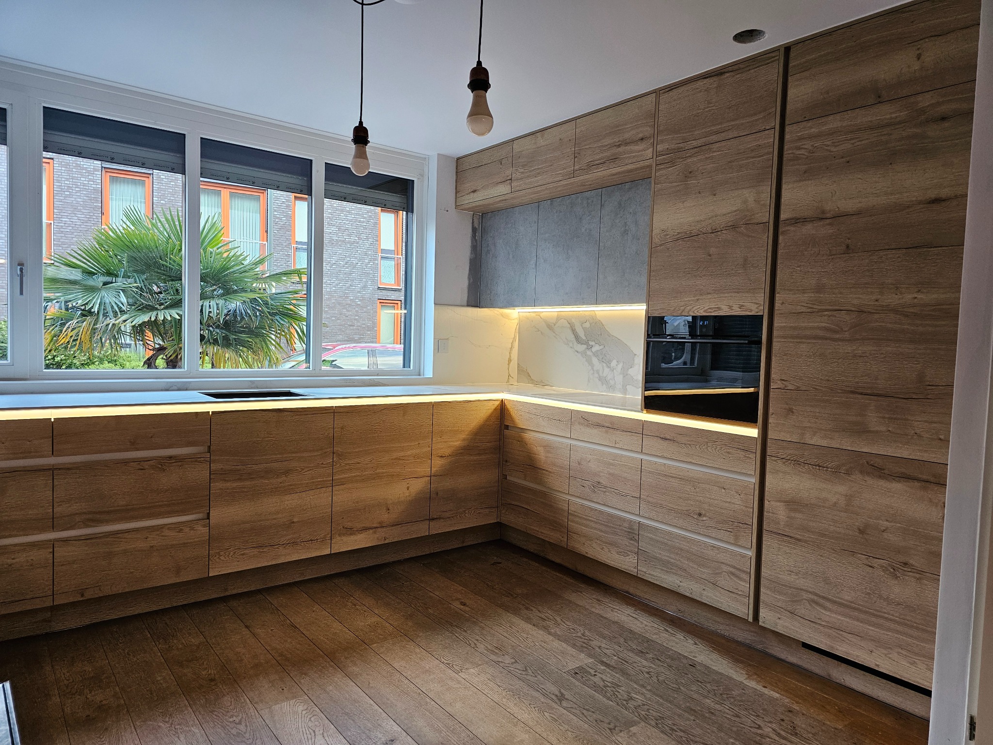 Nowoczesna kuchnia z drewnianymi frontami, oświetleniem LED pod blatem i widokiem na palmę za oknem.