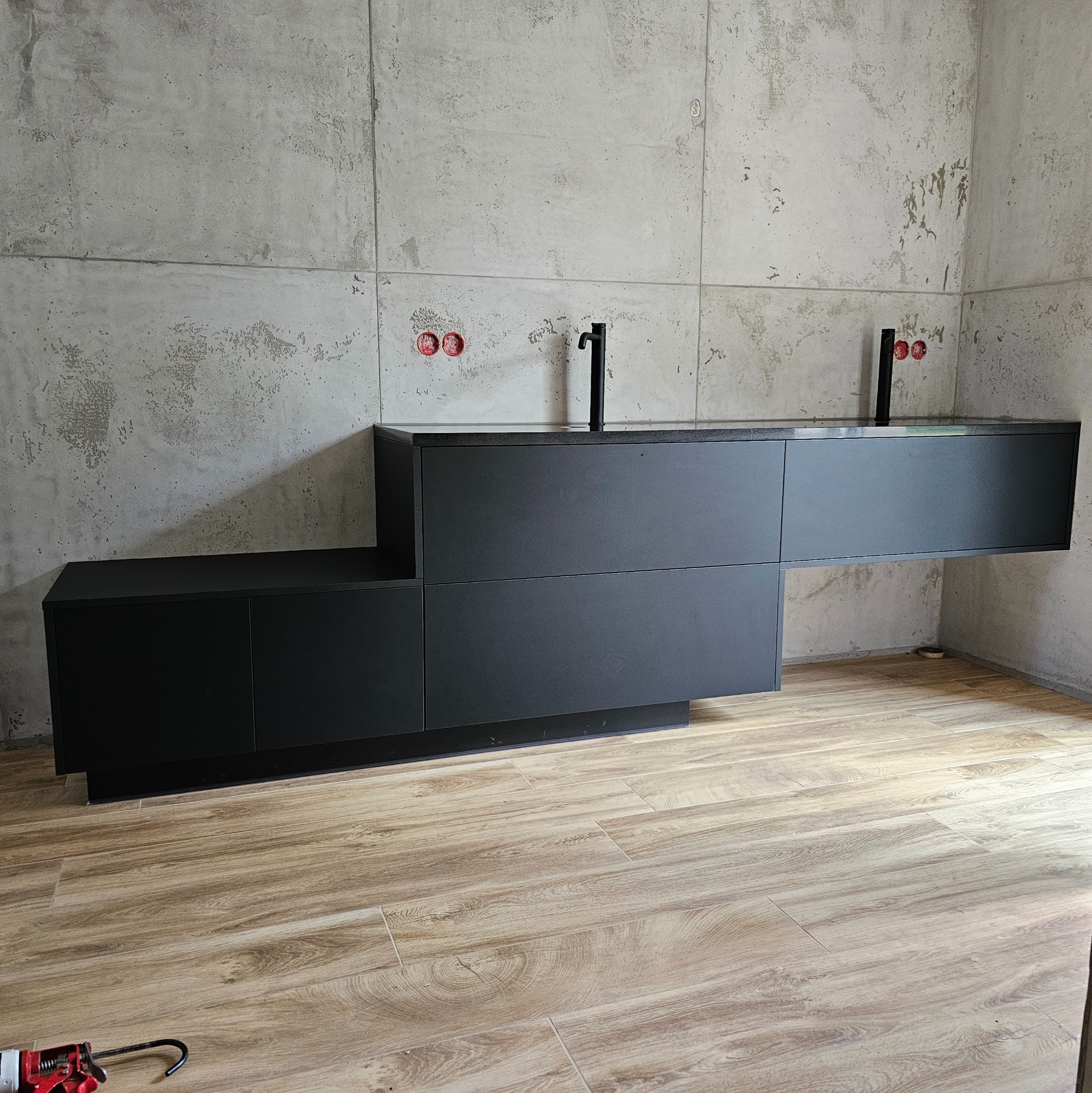 Czarna, nowoczesna szafka umywalkowa z dwiema umywalkami nablatowymi, zamontowana na tle surowej, betonowej ściany. Widoczne przyłącza hydrauliczne i narzędzie do silikonu na podłodze.