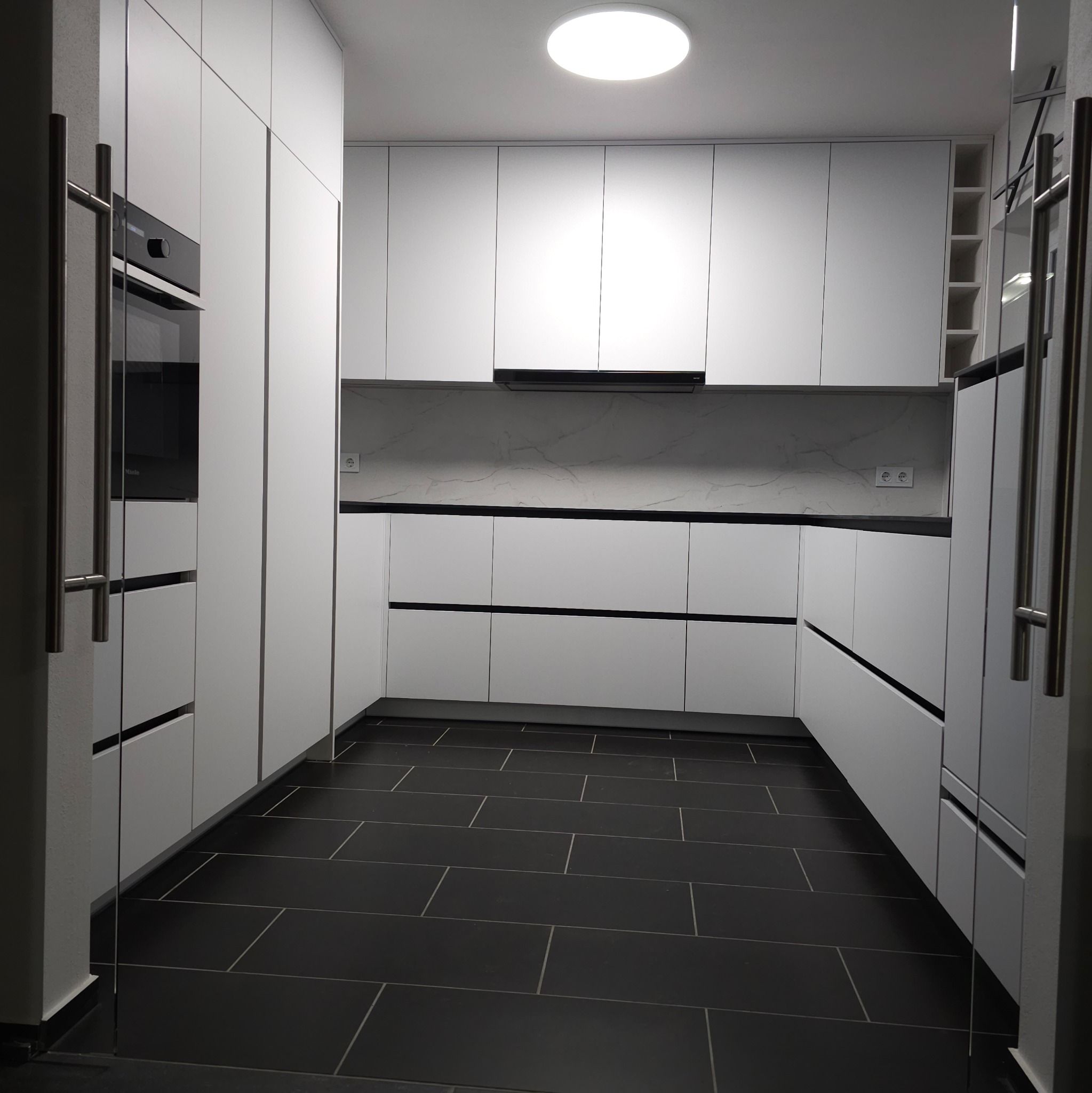 Nowoczesna, minimalistyczna kuchnia w bieli z czarną podłogą i blatem, oświetlona okrągłym plafonem, z widocznym piekarnikiem Miele i szklanymi drzwiami.