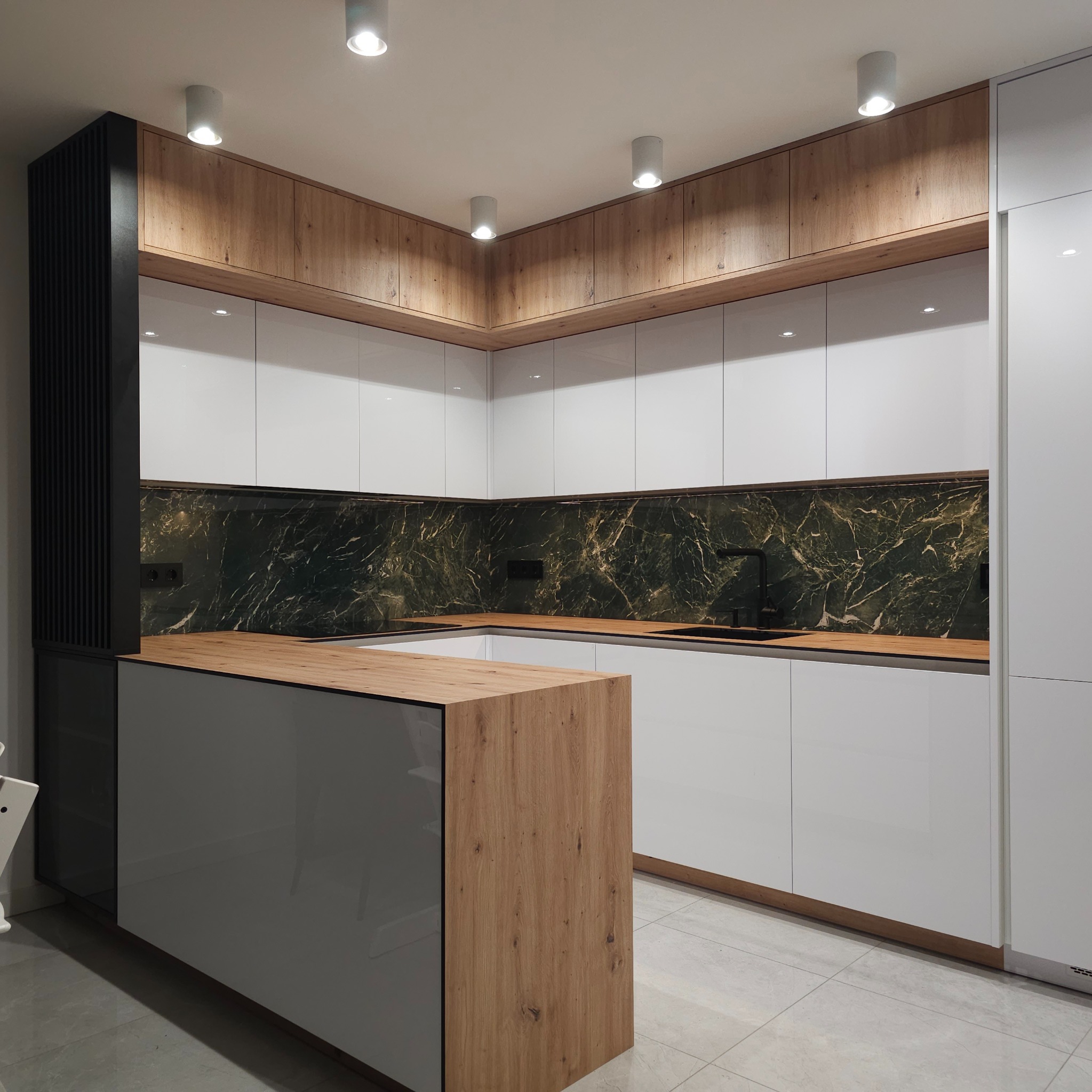 Nowoczesna kuchnia w kształcie litery U z białymi, połyskującymi frontami szafek, drewnianymi akcentami na górnych szafkach i wyspie, oraz ciemnozielonym panelem nad blatem imitującym marmur.