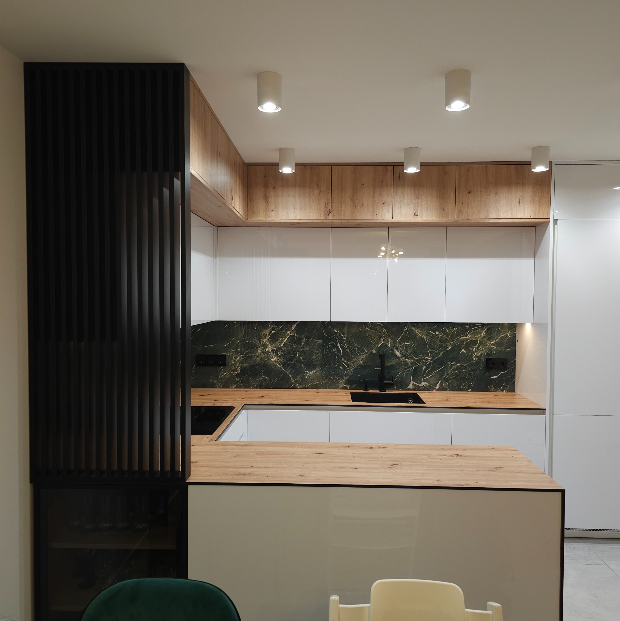 Nowoczesna kuchnia z białymi, lakierowanymi frontami, drewnianym blatem i panelem ściennym z motywem zielonego marmuru. Czarne akcenty w postaci baterii i listew. Element dekoracyjny z pionowych...