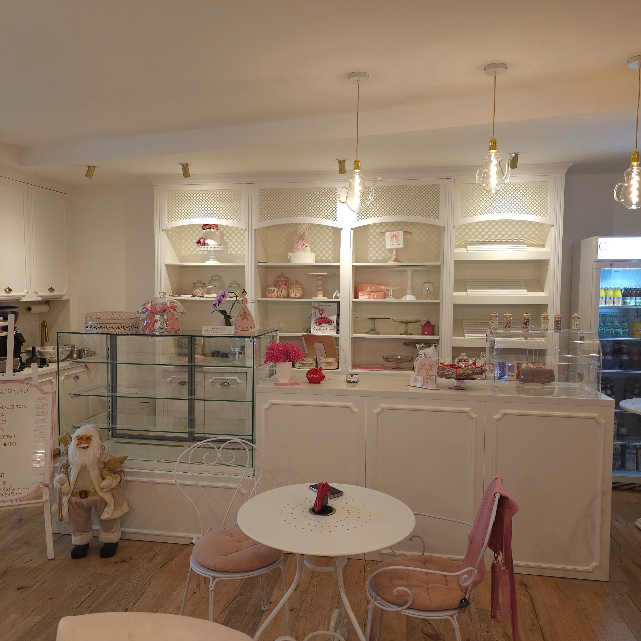 Jasne wnętrze cukierni z drewnianą podłogą, białymi meblami na wymiar z ażurowymi wstawkami, szklanymi witrynami z ciastami i dekoracjami, stolikiem z krzesłami oraz dekoracyjnymi lampami...