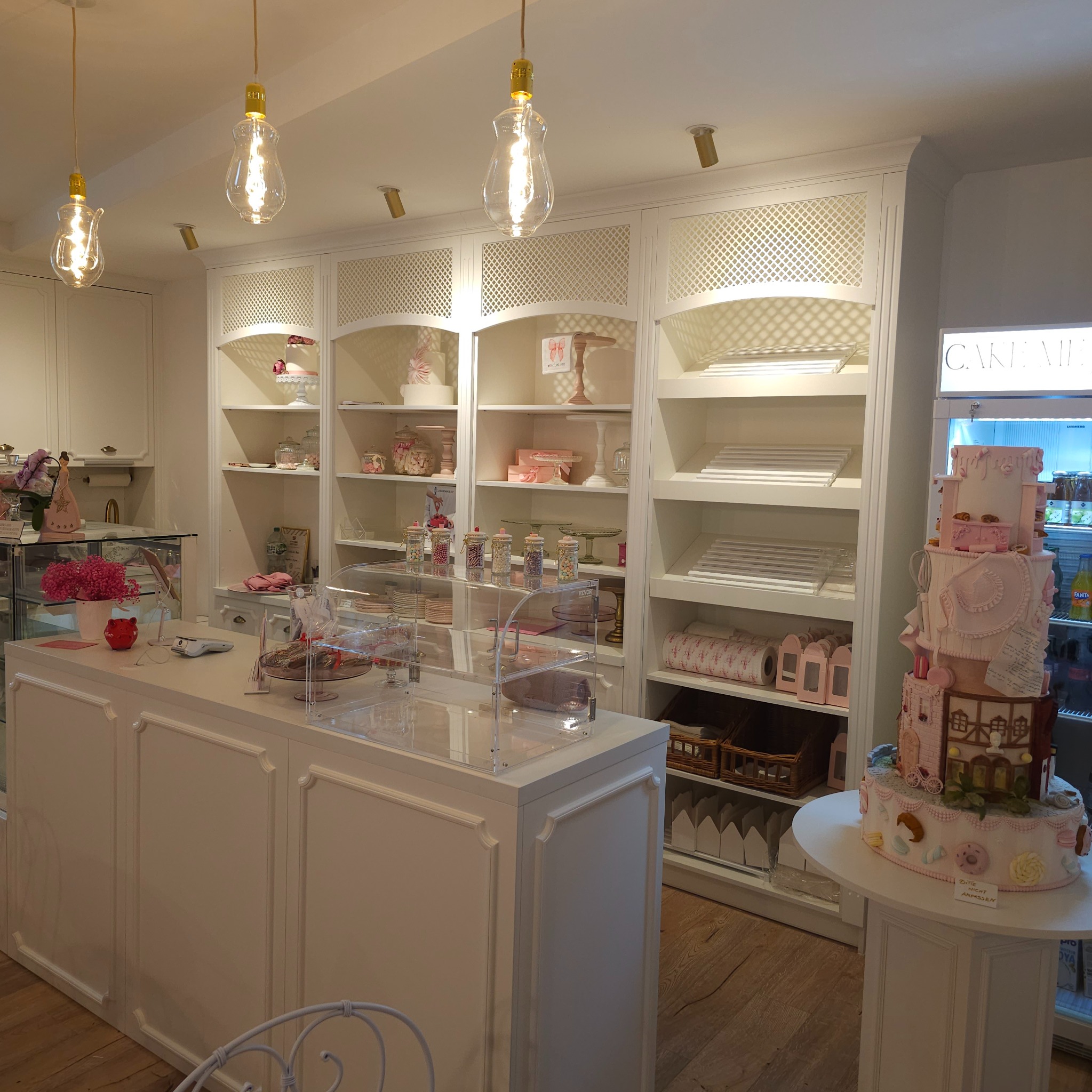 Wnętrze cukierni z białymi regałami wypełnionymi słodyczami i dekoracjami, na pierwszym planie lada i tort na postumencie.