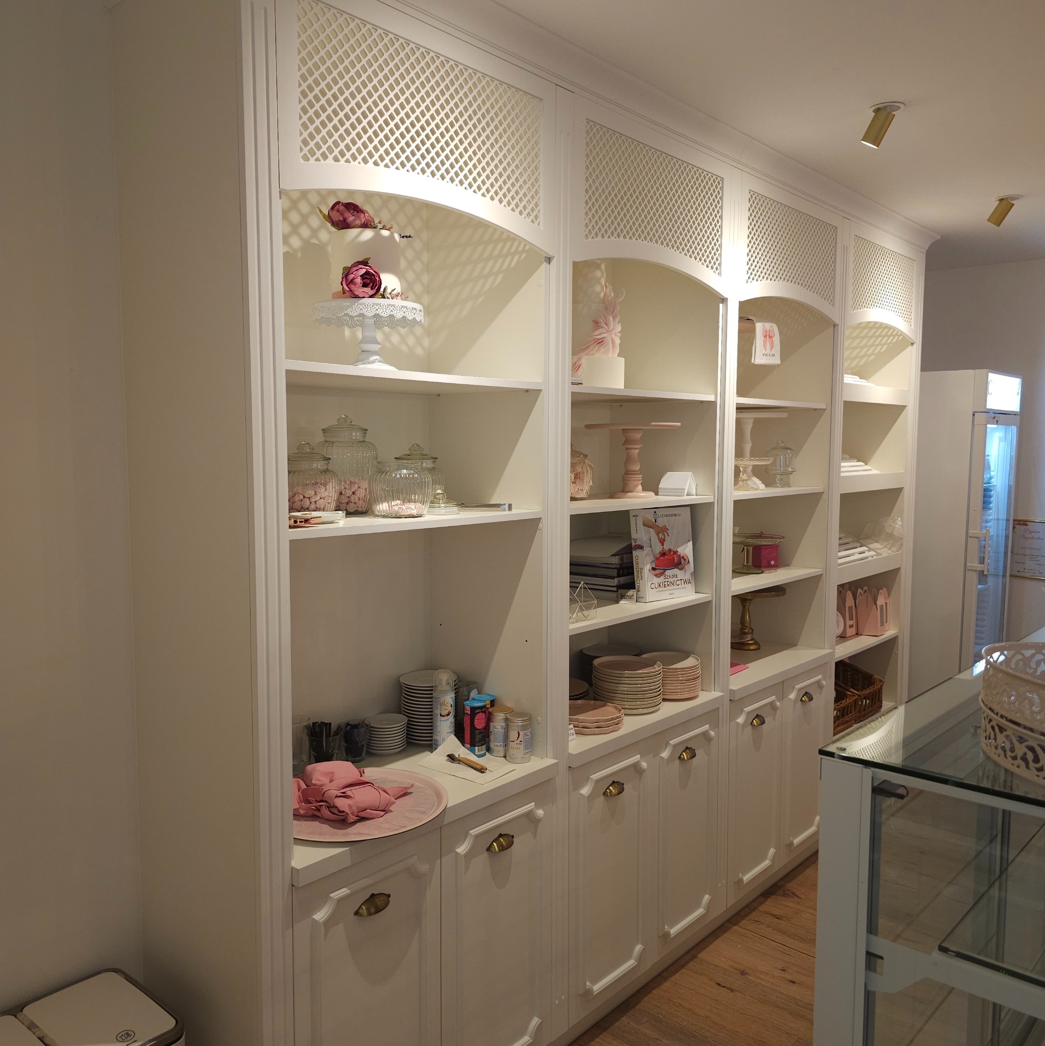 Biała zabudowa meblowa z ażurowymi frontami, ekspozycja cukiernicza z tortami, słodyczami i akcesoriami w pastelowych kolorach, drewniana podłoga.