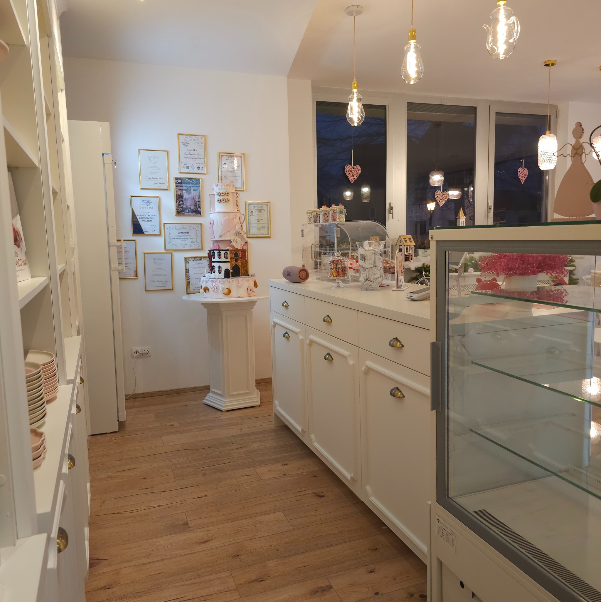 Wnętrze pracowni cukierniczej z tortem na postumencie, witryną chłodniczą i ścianą udekorowaną dyplomami.