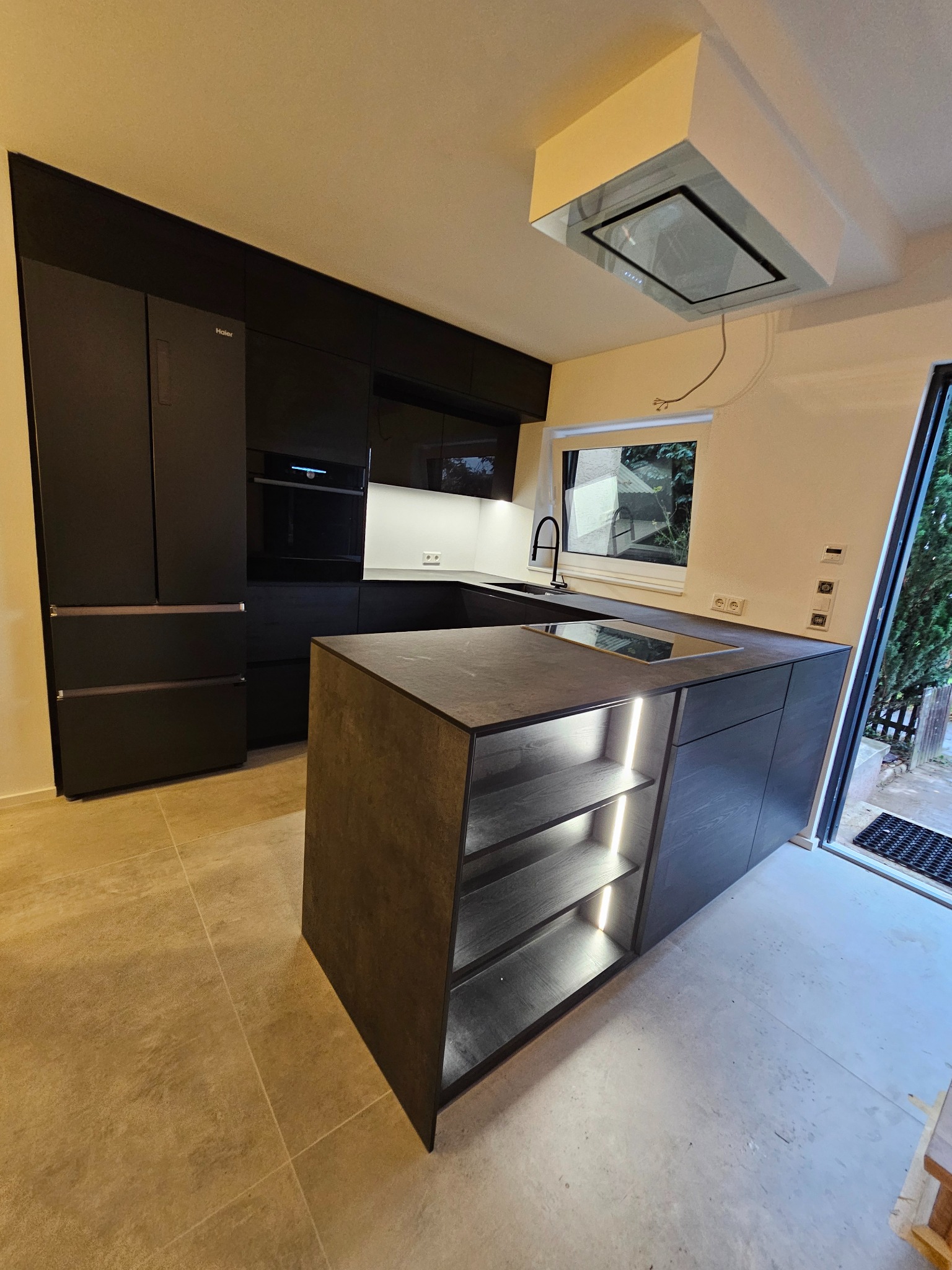 Nowoczesna, czarna kuchnia z wyspą oświetloną od dołu, widok na zabudowaną lodówkę i okap nad płytą grzewczą, jasne płytki na podłodze.