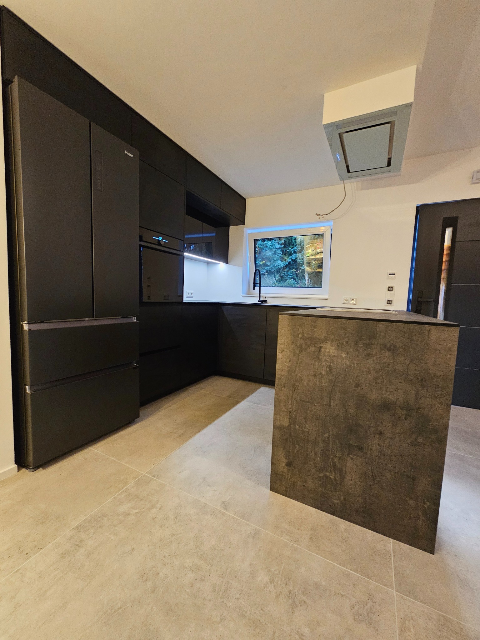 Nowoczesna kuchnia w odcieniach szarości i czerni z wyspą kuchenną o teksturze betonu, widok na okno z zielenią za nim i zabudowany sprzęt AGD marki Haier.