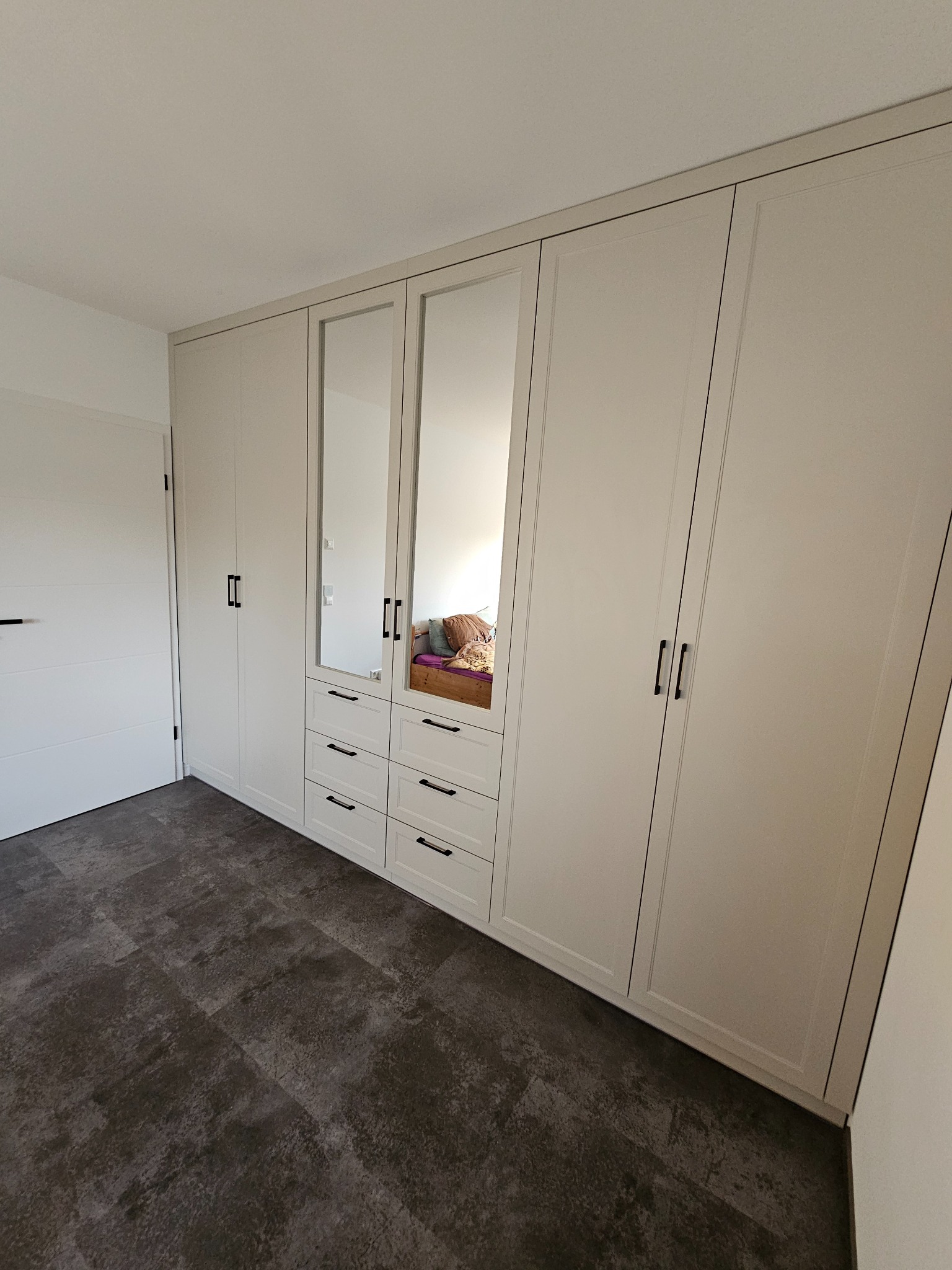 Biała szafa wnękowa z lustrami i czarnymi uchwytami, wkomponowana w jasne wnętrze z ciemną podłogą. Widoczny fragment pokoju z drzwiami.