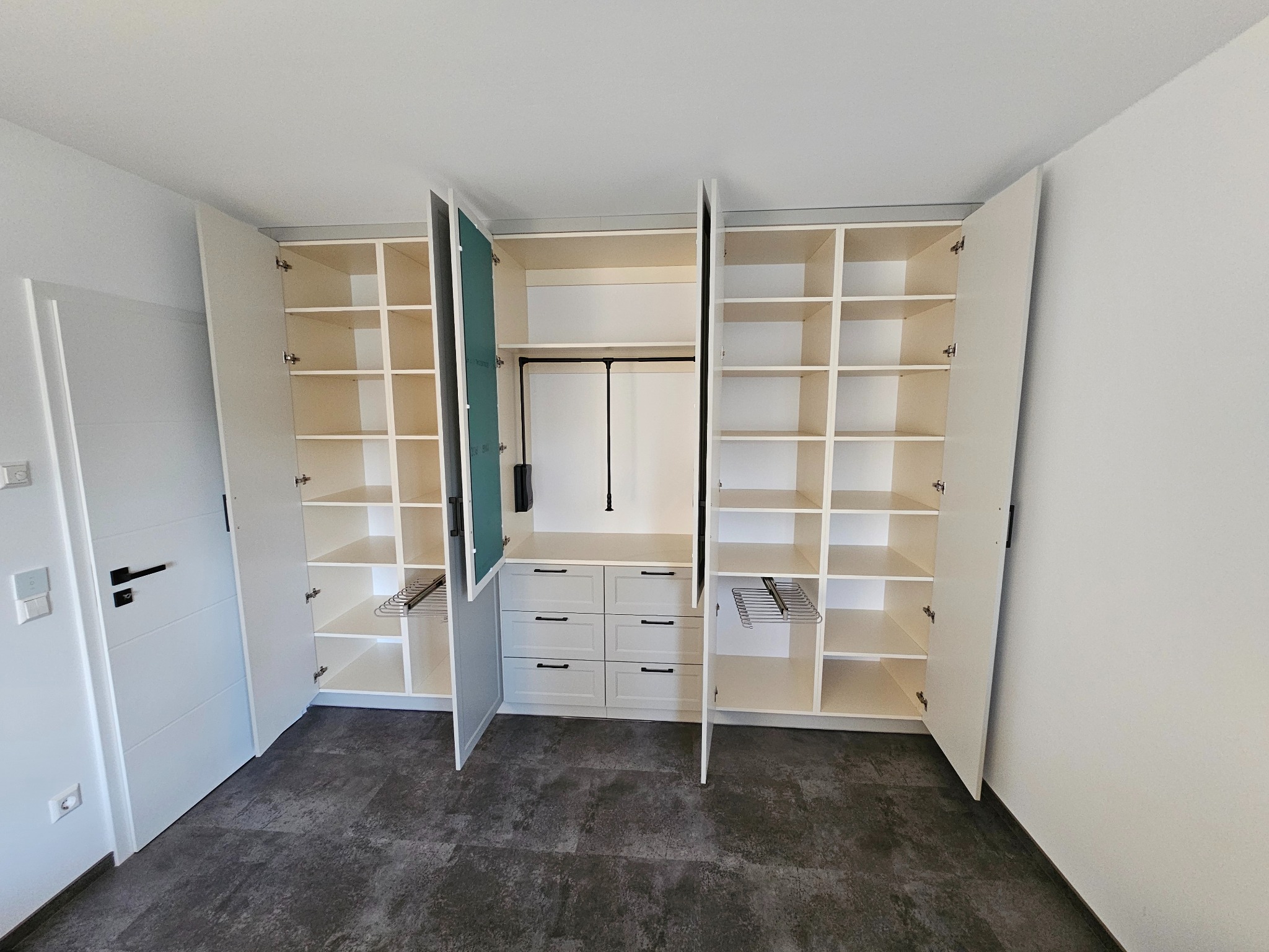 Biała, zabudowana szafa na wymiar z otwartymi drzwiami, widoczne półki i szuflady, metalowe wieszaki na spodnie, ciemna podłoga, minimalistyczny design.