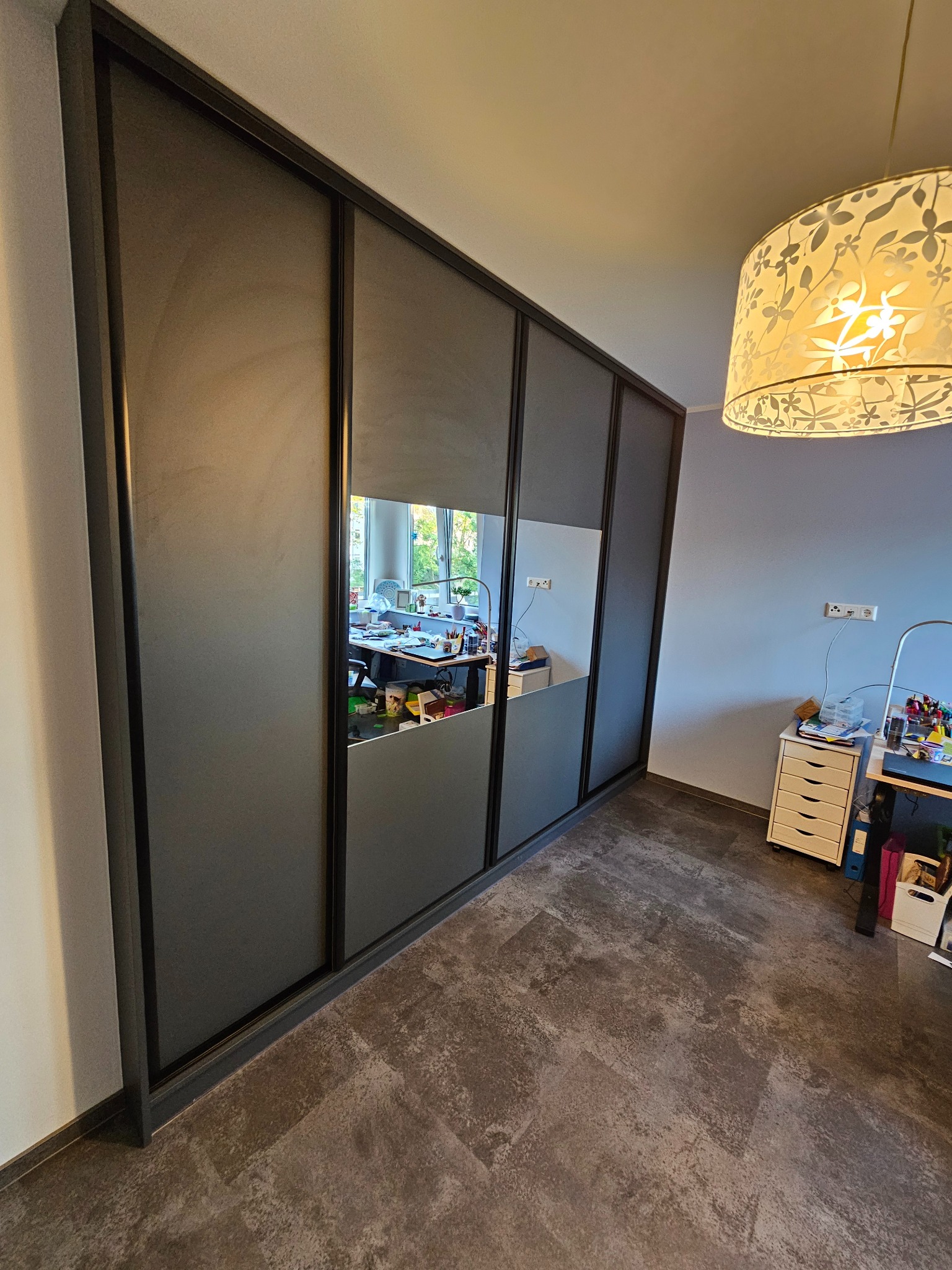 Szafa przesuwna z lustrem w szarej ramie, odbijająca biurko z akcesoriami, oświetlona lampą z motywem kwiatowym. Nowoczesny design, widok z perspektywy podłogi.