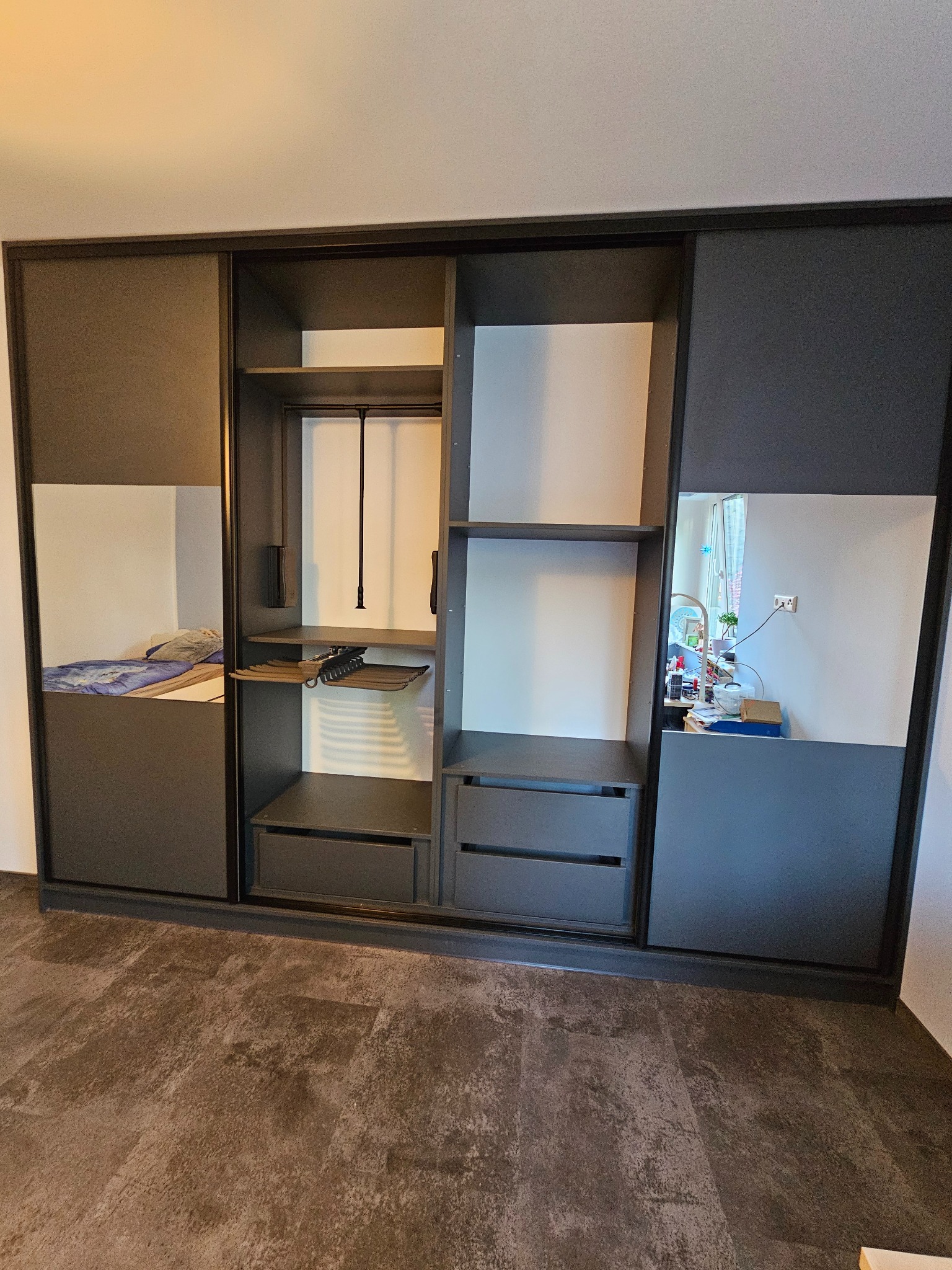 Szafa wnękowa z przesuwnymi drzwiami, wykończona w odcieniach szarości, z lustrzanymi panelami i widocznym wnętrzem z półkami i szufladami. Minimalistyczny design.