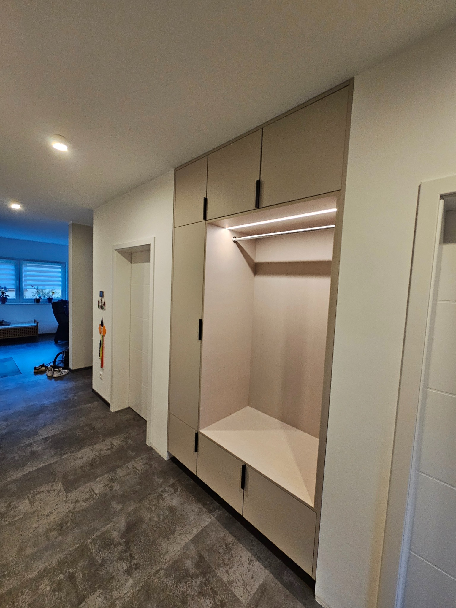 Beżowa zabudowa meblowa w przedpokoju z oświetleniem LED, szafkami i miejscem do siedzenia. Nowoczesny design, minimalistyczne uchwyty, szara podłoga.