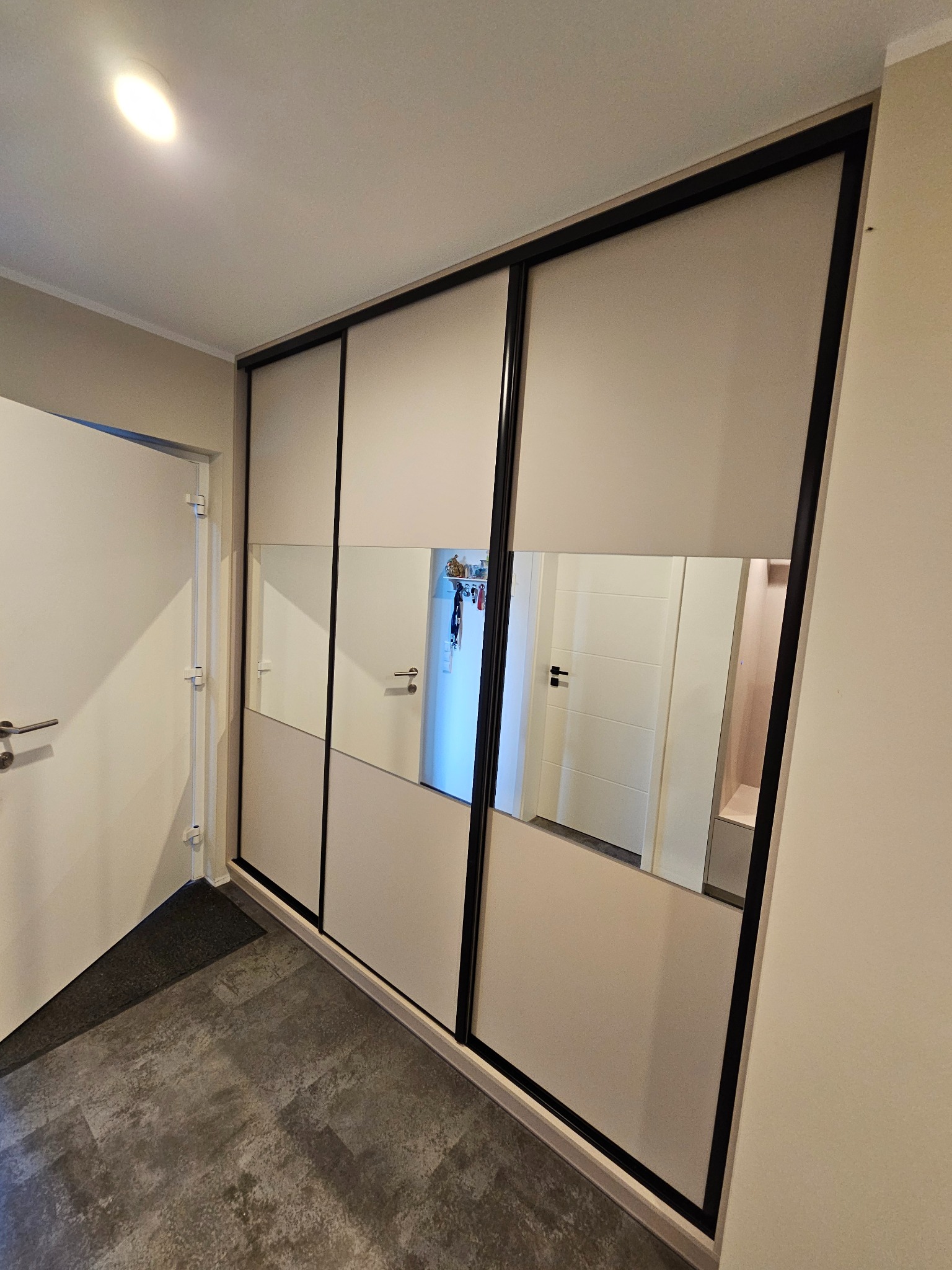 Biała szafa przesuwna z czarnymi obramowaniami i lustrami, w których odbija się wnętrze mieszkania, na tle szarej podłogi i białych ścian.