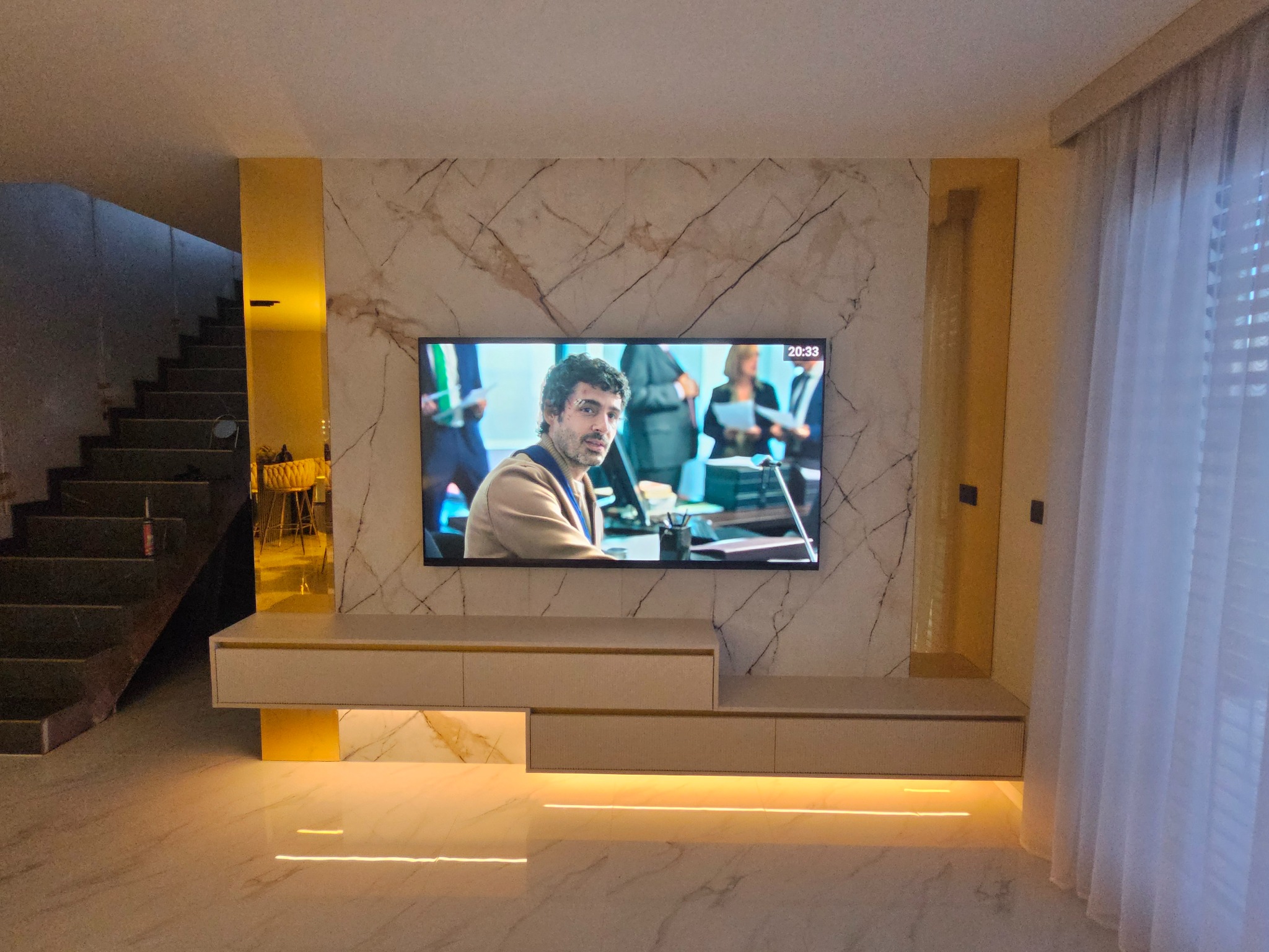 Nowoczesna szafka RTV na wymiar z podświetleniem LED, dopasowana do marmurowej ściany i podłogi. Telewizor zawieszony nad szafką. Minimalistyczny design.