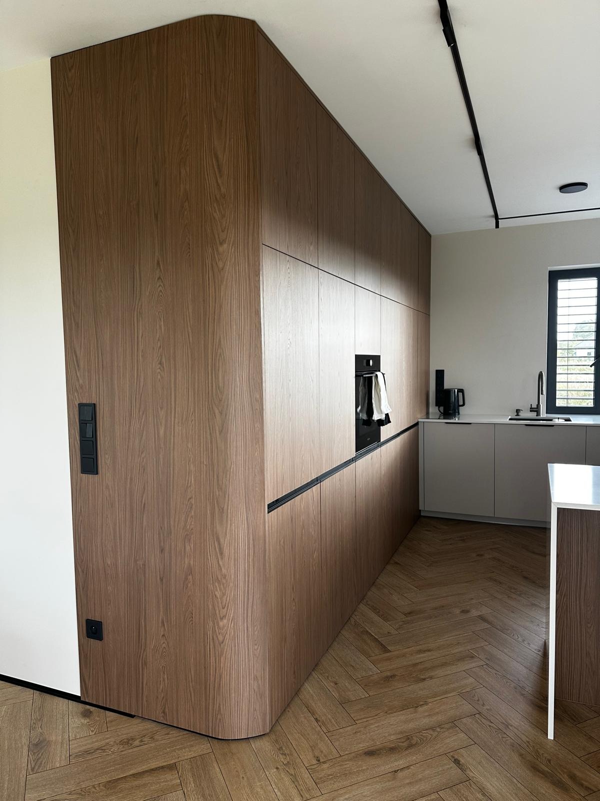 Nowoczesna kuchnia z drewnianymi frontami, zabudowana od podłogi do sufitu, z zaokrąglonym narożnikiem i czarnymi akcentami, na podłodze parkiet w jodełkę.