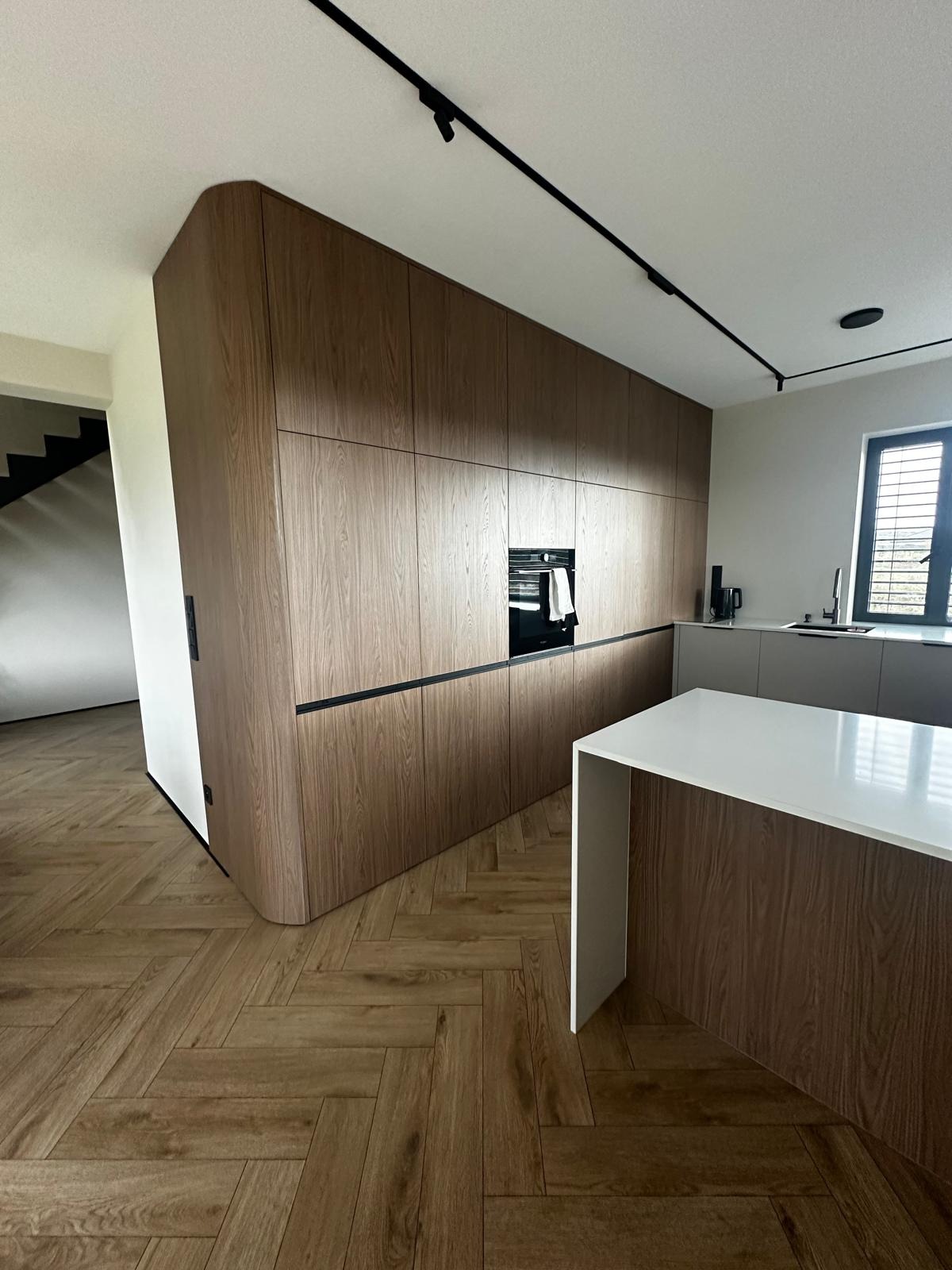 Nowoczesna kuchnia z drewnianymi frontami, czarnymi uchwytami i oświetleniem szynowym. Wyspa kuchenna z białym blatem. Parkiet w jodełkę. Minimalistyczny design.