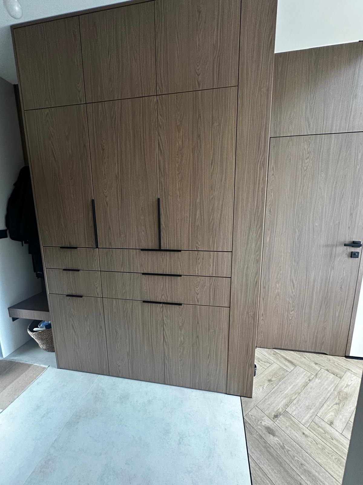 Szafa wnękowa z drewnopodobnymi frontami i czarnymi uchwytami, dopasowana kolorystycznie do drzwi, z widocznym fragmentem podłogi w jodełkę.