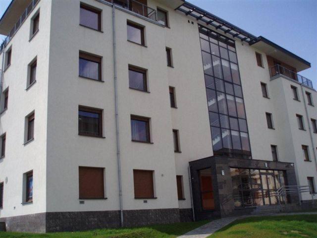 Elewacja budynku wielorodzinnego z widocznymi oknami z brązowymi roletami zewnętrznymi i szklaną fasadą wejścia.