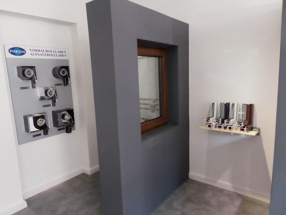 Ekspozycja okna z drewnianą ramą w szarej wnęce, obok plansza z roletami i próbki profili okiennych na półce. Prezentacja stolarki okiennej w salonie.