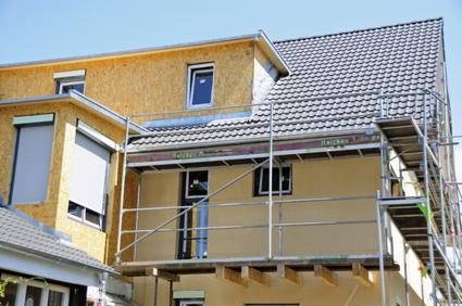 Budynek w trakcie prac elewacyjnych, z oknami i częściowo opuszczoną roletą, widoczny z rusztowania. Jedna część budynku z ociepleniem, druga z tynkiem.