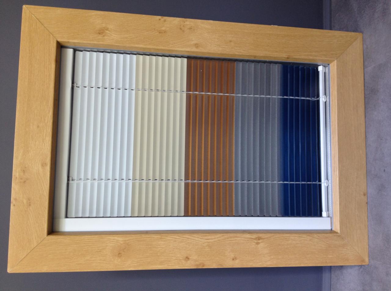 Drewniana rama okienna z ekspozycją rolet plisowanych w różnych kolorach: biel, beż, brąz, szary i granat. Prezentacja próbek kolorystycznych rolet.