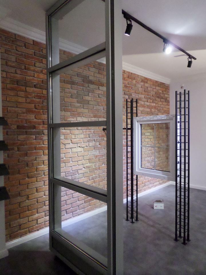 Wnętrze z ekspozycją okien PVC na tle ściany z cegły. Widoczne ramy okienne w kolorze białym i szarym. Nowoczesne oświetlenie sufitowe.