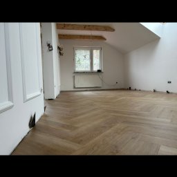 Wysoki standart wykończenia wnętrz ,łazienek,remonty - Jasny parkiet ułożony w jodełkę francuską w przestronnym pokoju na poddaszu. Widoczne białe ściany, okno z grzejnikiem i fragment drewnianej konstrukcji dachu.