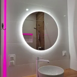 Nowoczesna łazienka z okrągłym lustrem LED, białą umywalką nablatową i blatem z drewna. Widoczna kabina prysznicowa i akcenty w postaci różowego oświetlenia.