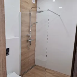 Nowoczesna łazienka z kabiną typu walk-in, wykończona białymi płytkami i panelami imitującymi drewno. Widoczny odpływ liniowy i minimalistyczna armatura.