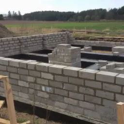 Wznoszenie fundamentów z bloczków betonowych, widoczne izolacja przeciwwilgociowa, stos bloczków i hałda ziemi w tle.