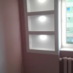 Różowy pokój z wnęką z płyt karton gips z trzema półkami oświetlonymi od góry, obok okna z widokiem na blok mieszkalny.