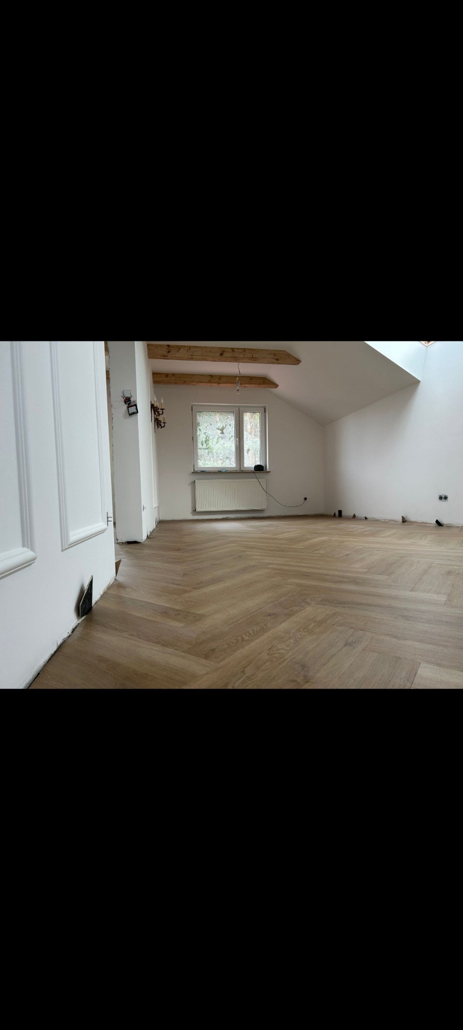 Jasny parkiet ułożony w jodełkę francuską w przestronnym pokoju na poddaszu. Widoczne białe ściany, okno z grzejnikiem i fragment drewnianej konstrukcji dachu.