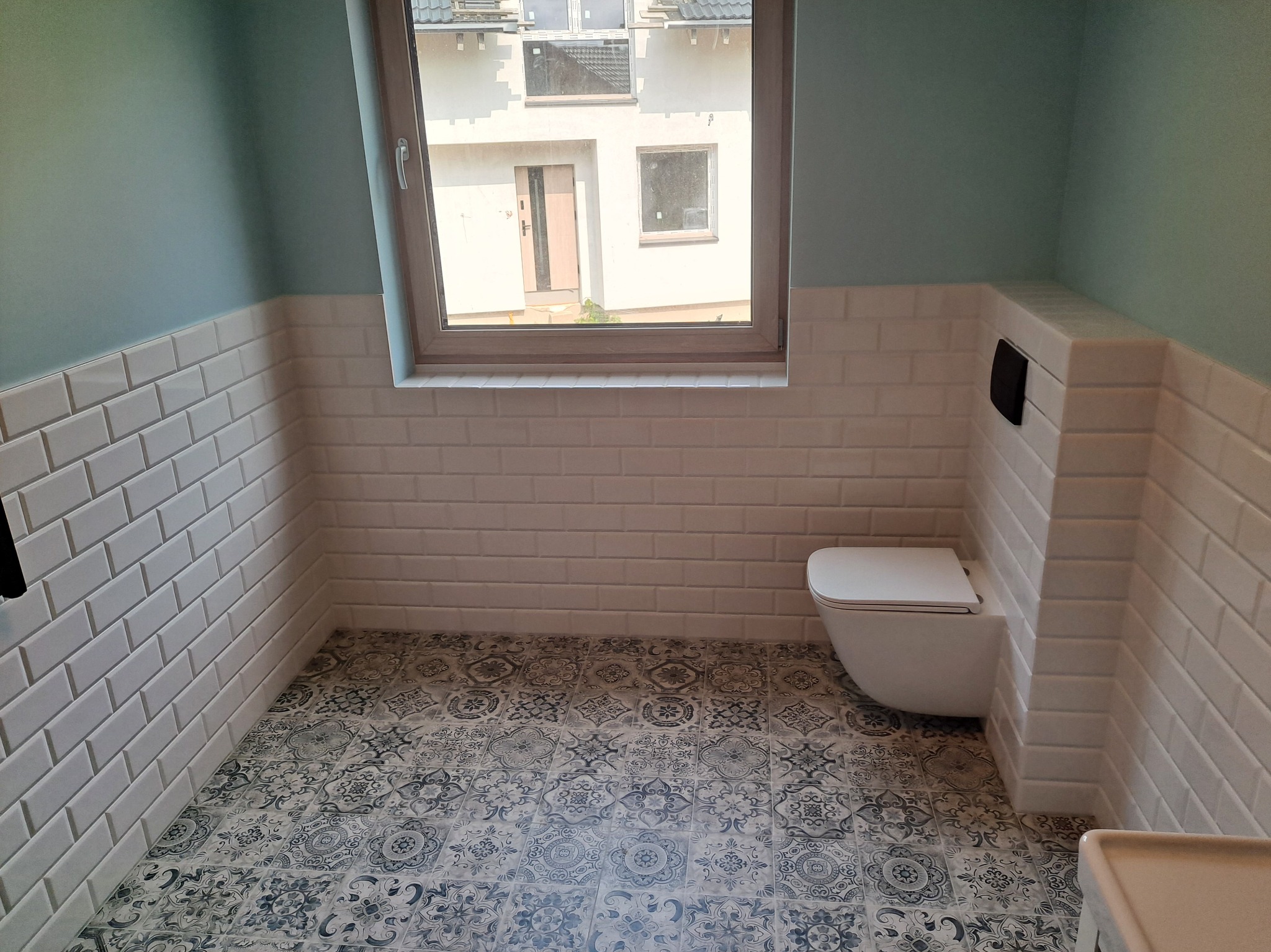 Wykończona łazienka w stylu retro: białe kafelki do połowy ściany, wzorzysta podłoga i podwieszana toaleta. Okno z widokiem na budynek. Odświeżony minimalizm.