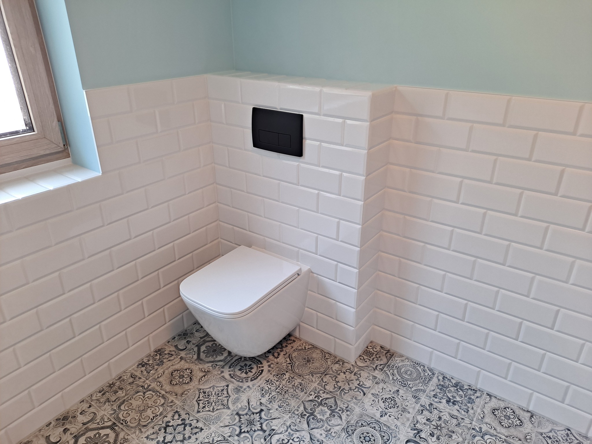 Biała, mała łazienka z podwieszaną toaletą, wzorzystą podłogą i płytkami typu metro na ścianach. Ciemny przycisk spłukiwania. Okno w rogu.