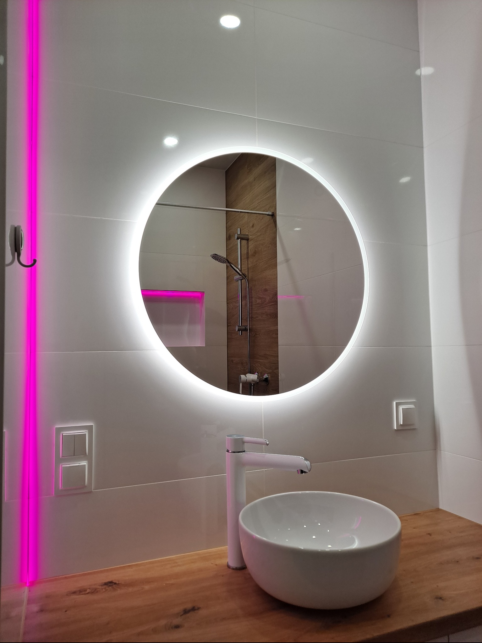 Nowoczesna łazienka z okrągłym lustrem LED, białą umywalką nablatową i blatem z drewna. Widoczna kabina prysznicowa i akcenty w postaci różowego oświetlenia.