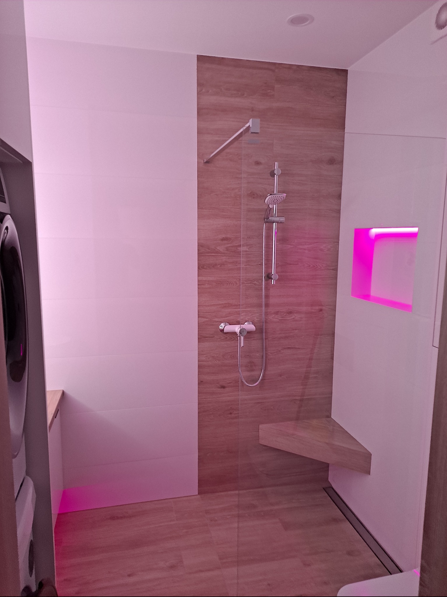 Nowoczesna kabina prysznicowa z drewnopodobnymi płytkami, deszczownicą i półką. W tle pralka i podświetlane wnęki, tworzące unikalny klimat w łazience.