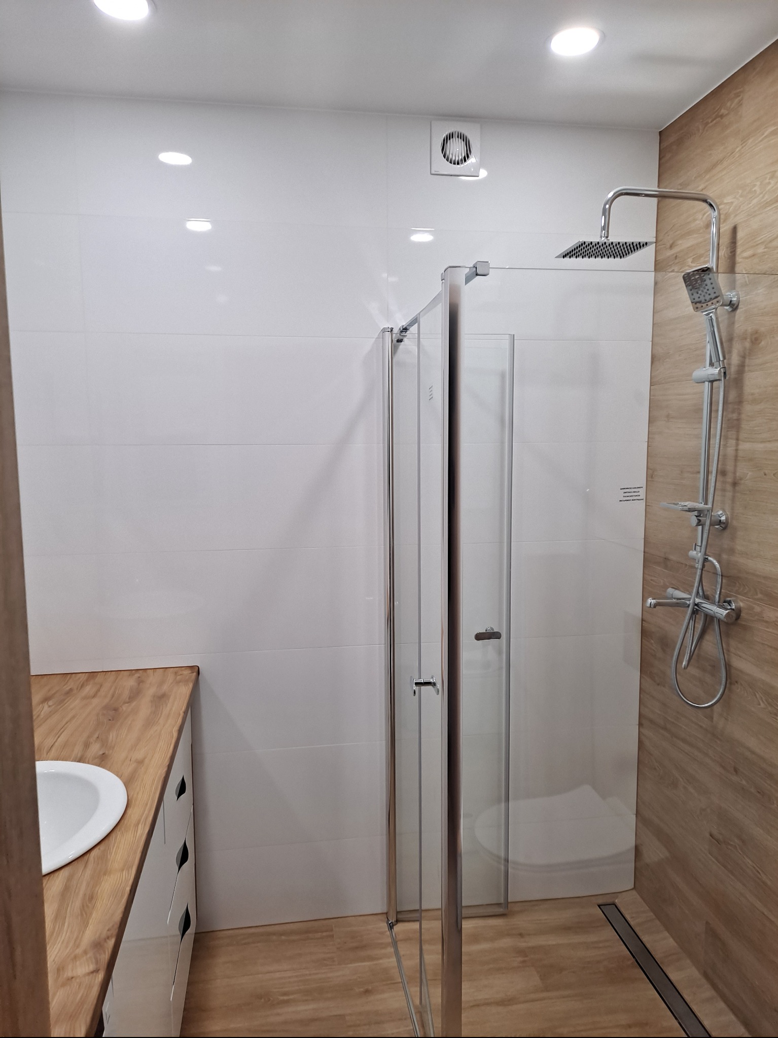 Nowoczesna łazienka z kabiną prysznicową typu walk-in, wykończona białymi i drewnopodobnymi płytkami, podkreślona punktowym oświetleniem sufitowym i minimalistyczną armaturą.