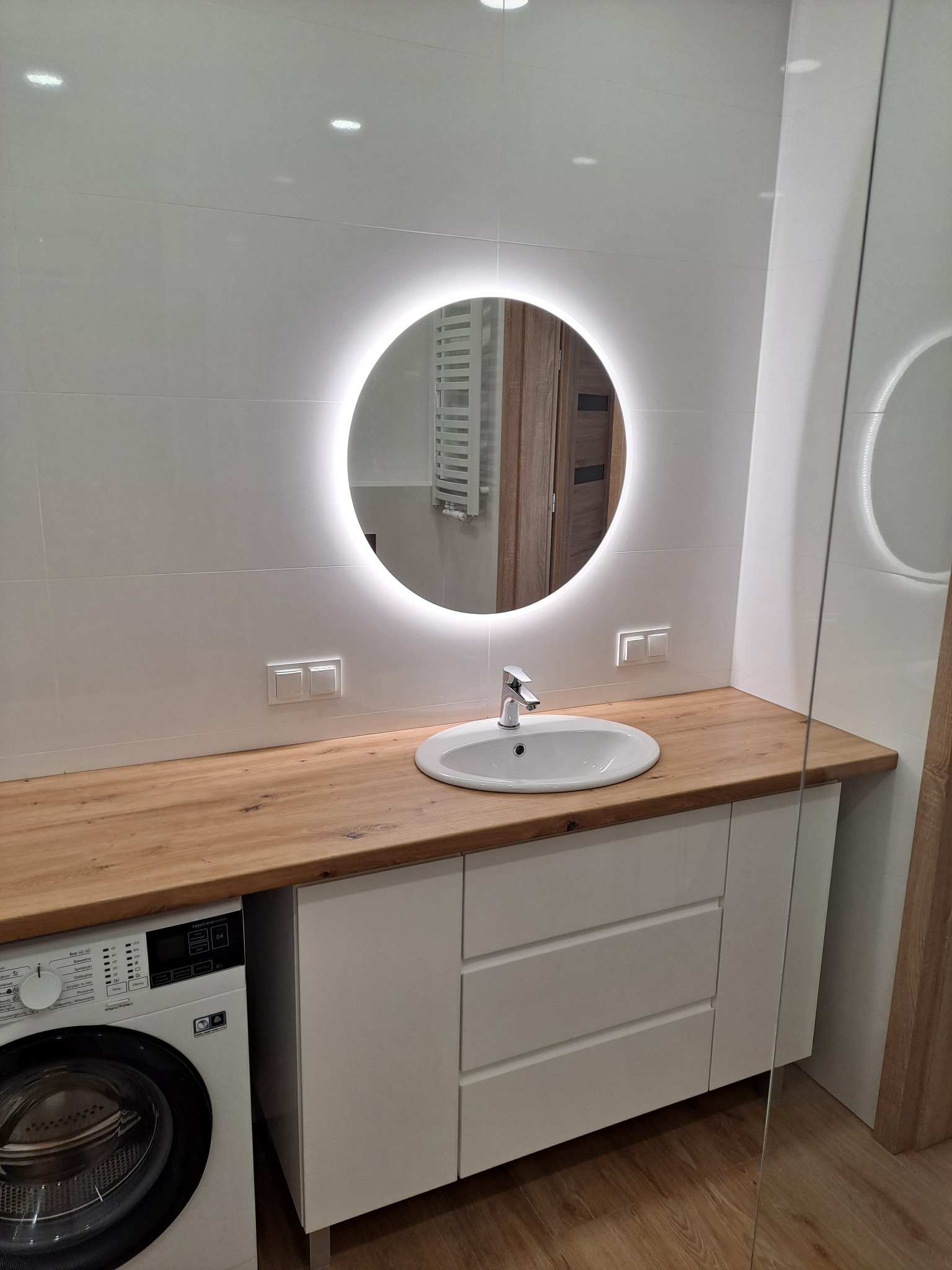 Jasna łazienka z okrągłym lustrem LED, drewnianym blatem i białą umywalką. Obok pralka. Białe płytki na ścianach i drewniana podłoga.